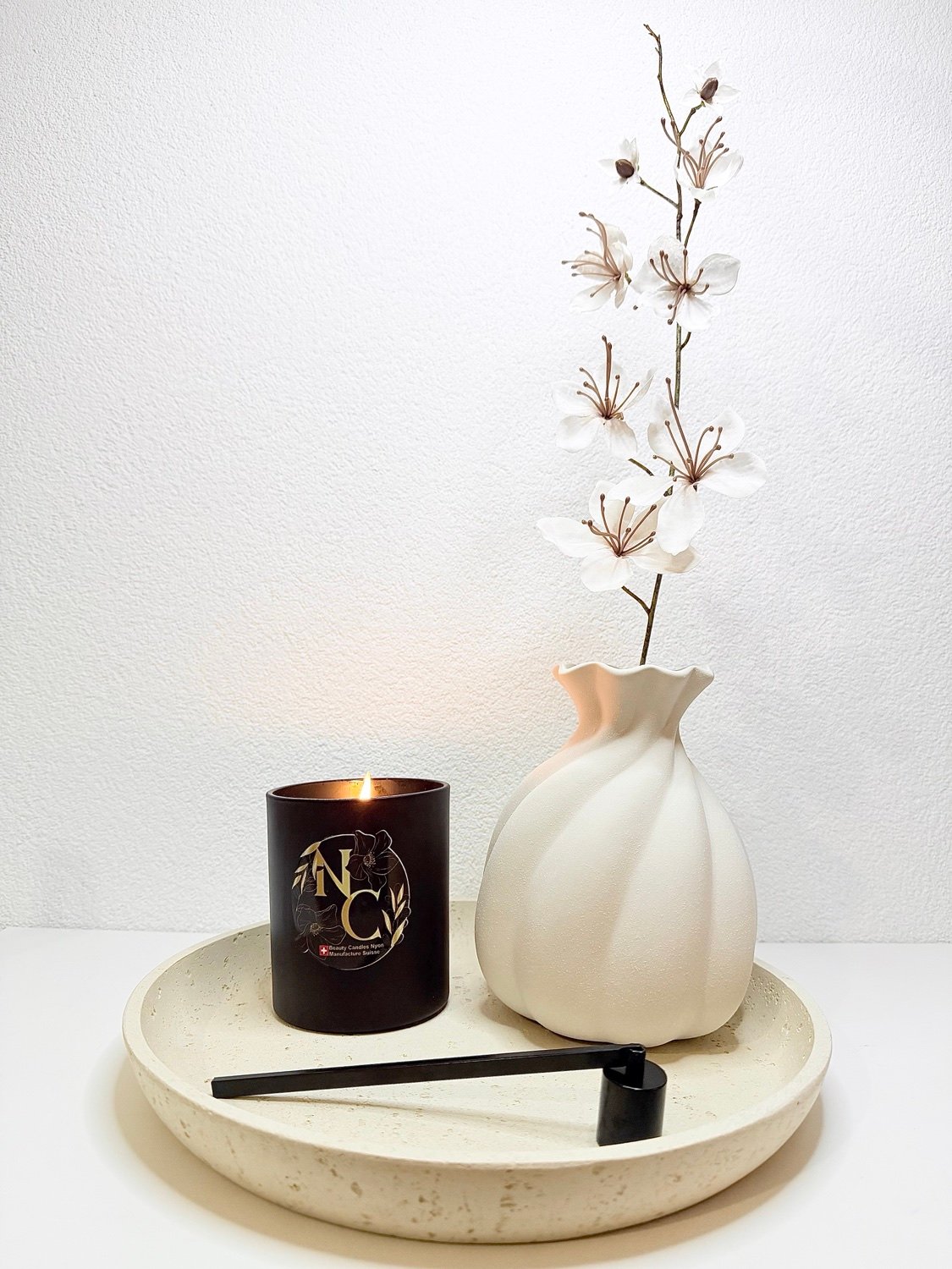 Bougie NC Candles allumée sur un plateau, à côté d'un vase blanc et d'une branche fleurie, avec l'éteignoir de la maison
